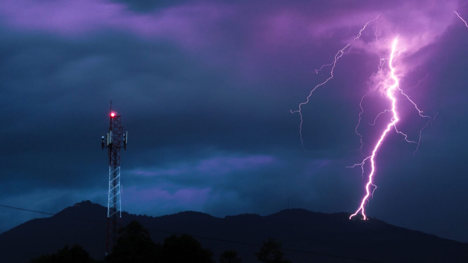 Vivid lightning bolt illuminates the night sky above a mountain and telecom tower.